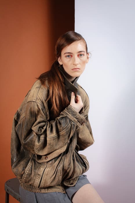 Portrait of a young woman with auburn hair, wearing a brown jacket and sitting on a metal stool, posing against a split background.