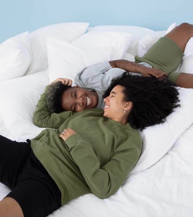 Two smiling people lying on a white bed with pillows. One person wears an olive sweatshirt. The other wears a gray top and olive shorts.