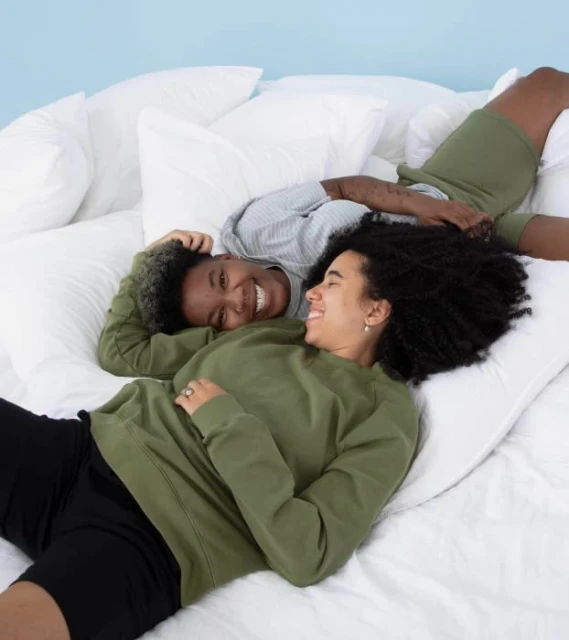 Two smiling people lying on a white bed with pillows. One person wears an olive sweatshirt. The other wears a gray top and olive shorts.