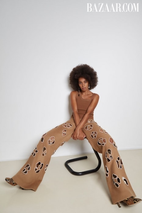 A woman with an afro poses in wide-leg, tan pants with cut-out designs, a matching tank top, and snakeskin-print heels against a white wall.