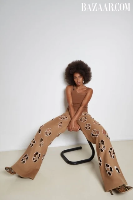 A woman with an afro poses in wide-leg, tan pants with cut-out designs, a matching tank top, and snakeskin-print heels against a white wall.