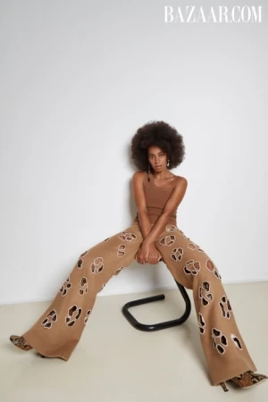 A woman with an afro poses in wide-leg, tan pants with cut-out designs, a matching tank top, and snakeskin-print heels against a white wall.