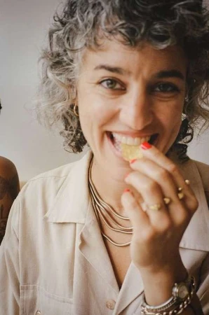 Close-up portrait of a smiling woman with curly grey-streaked hair eating a chip, wearing a beige shirt and gold layered necklaces.