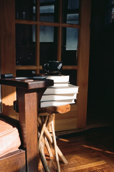 A stack of books sits on a wooden side table with a coffee mug next to a wooden door, creating a cozy, warm, and inviting scene.