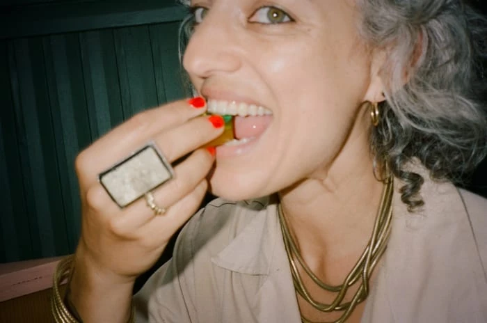 Close-up of a woman with gray hair eating candy; she wears gold jewelry and red nail polish against a dark-green background.