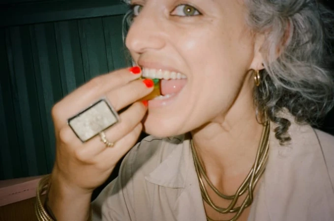 Close-up of a woman with gray hair eating candy; she wears gold jewelry and red nail polish against a dark-green background.