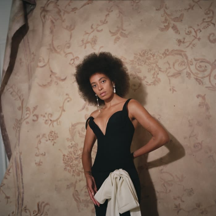 A woman with an afro hairstyle poses in a black off-the-shoulder dress with a cream-colored bow against a patterned backdrop.