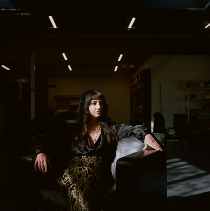 Woman with long dark hair sits on a black leather chair in an indoor space with overhead fluorescent lighting and shelves in the background.