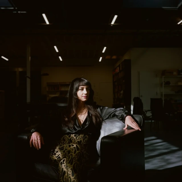 Woman with long dark hair sits on a black leather chair in an indoor space with overhead fluorescent lighting and shelves in the background.
