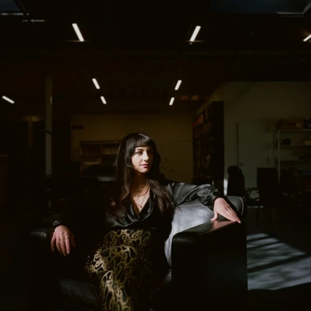 Woman with long dark hair sits on a black leather chair in an indoor space with overhead fluorescent lighting and shelves in the background.