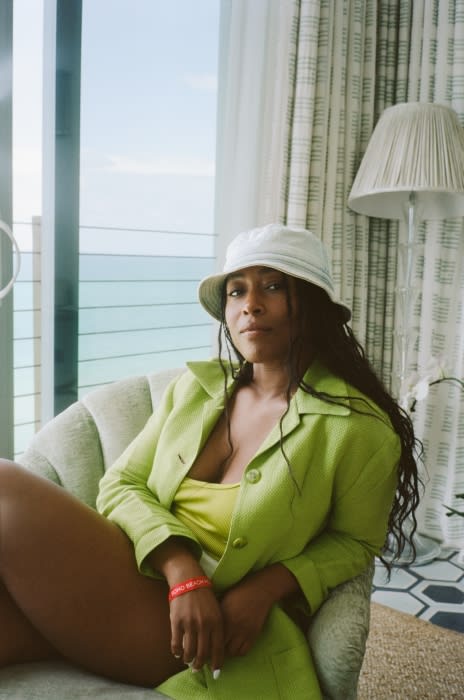 Adult female with long dark hair in a lime green suit jacket sitting in a light green chair with an ocean view from a high-rise apartment.