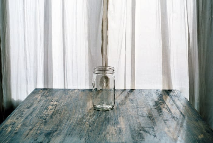Empty glass mason jar sitting on a blue wooden table, with sheer white curtains hanging in the background.