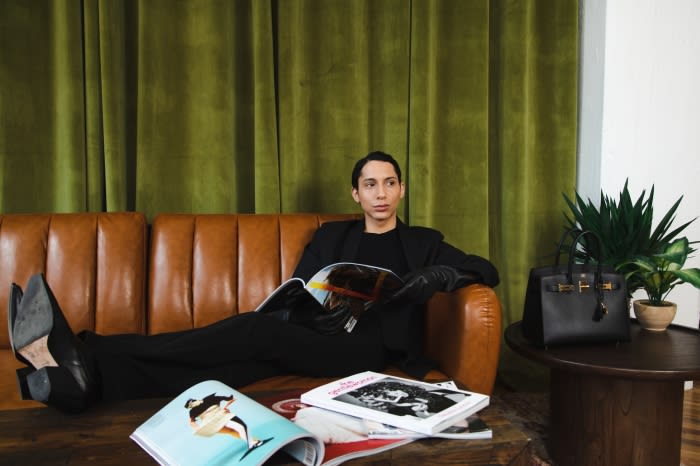 Person in black attire seated on a brown leather sofa holding an open magazine with green velvet curtains behind
