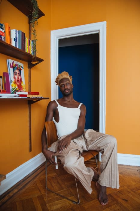 An African American adult male poses indoors, sitting in a chair in front of a doorway in a room with bright-orange walls and wood floors.