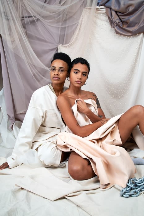 Two women pose indoors against a backdrop of draped fabrics, one wearing glasses and a jacket, the other draped in a light fabric.