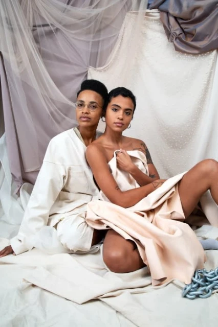 Two women pose indoors against a backdrop of draped fabrics, one wearing glasses and a jacket, the other draped in a light fabric.