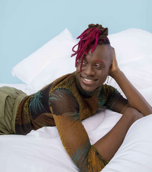 Smiling African-descent adult with dreadlocks and a sheer palm-print top, reclining on a white bed against a pale blue wall.