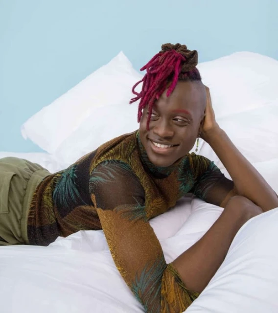 Smiling African-descent adult with dreadlocks and a sheer palm-print top, reclining on a white bed against a pale blue wall.