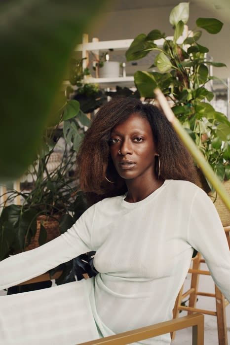 Portrait of a Black woman in a light green outfit surrounded by green plants and natural lighting in an indoor setting.