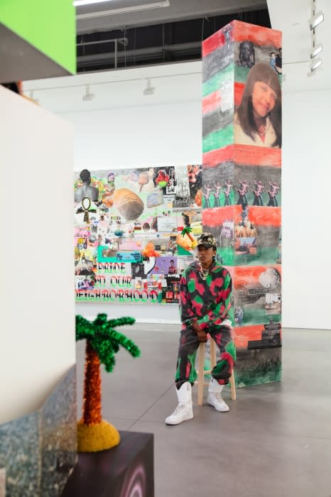 Man in colorful outfit sits on a stool in an art gallery with paintings and installations, creating a vibrant and contemporary scene.