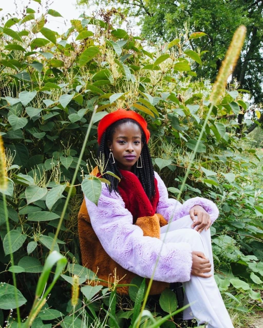 A woman with dark skin and braided hair wearing a red beret, colorful coat, and white pants is sitting outdoors among green leafy plants.