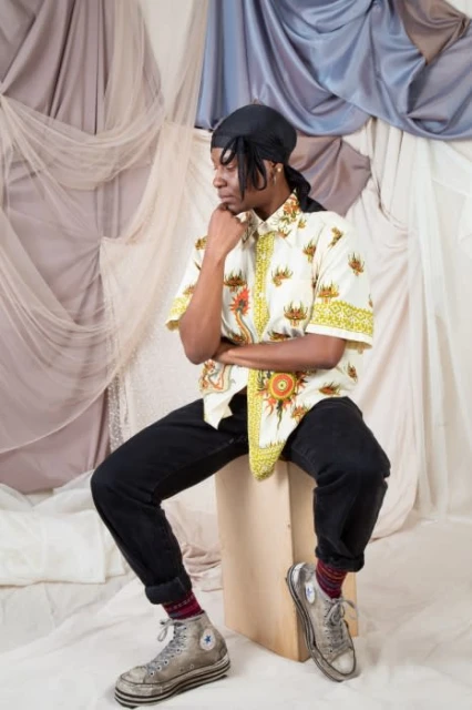 Young adult in patterned shirt and jeans, sitting on a wooden cube, with neutral backdrop and old Converse sneakers, in a casual pose.