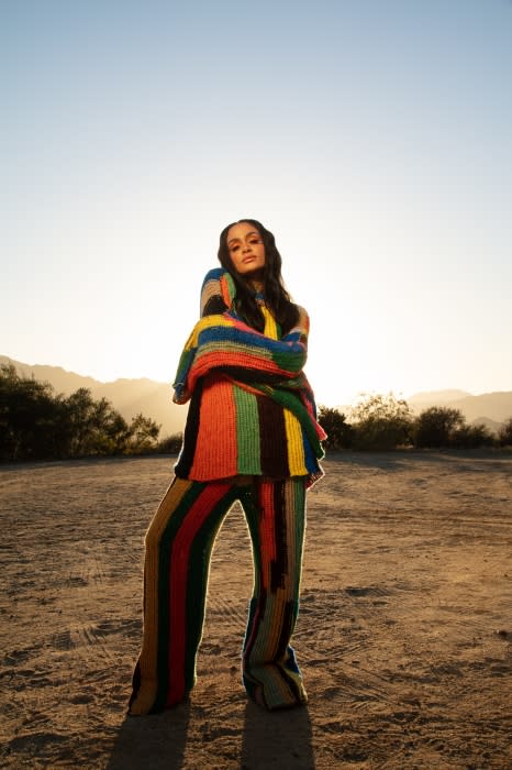 Woman in colorful knitwear poses in a desert landscape at sunset, wearing striped sweater and pants, with mountains in background.