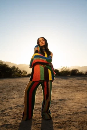 Woman in colorful knitwear poses in a desert landscape at sunset, wearing striped sweater and pants, with mountains in background.