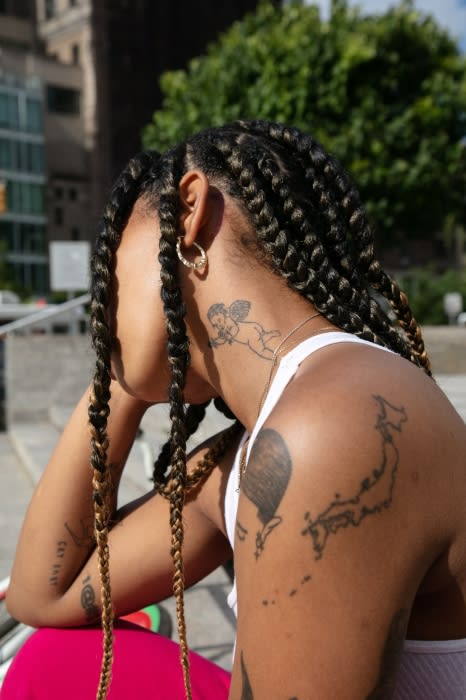 Woman with braided hair and tattoos poses outside against a city backdrop, showcasing a fashionable and artistic aesthetic.