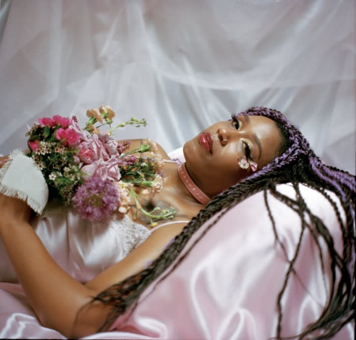 Woman with braided purple hair, laying in a pink satin bed, adorned with flowers and light makeup for a dreamy, soft aesthetic.