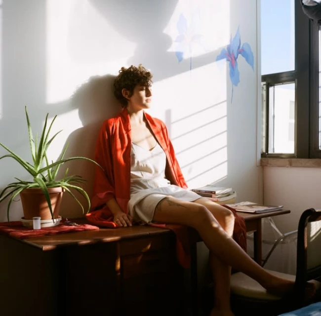 Woman with short curly hair sits barefoot on a table in a brightly lit interior. She wears a slip dress and red robe.