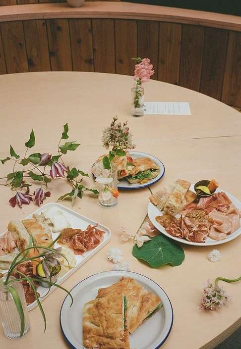 Overhead view of a variety of sandwiches and charcuterie on plates and platters, flowers in vases on a round wooden table.
