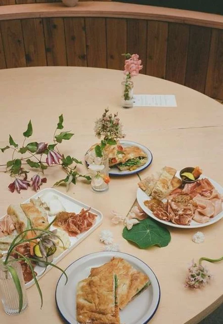 Overhead view of a variety of sandwiches and charcuterie on plates and platters, flowers in vases on a round wooden table.