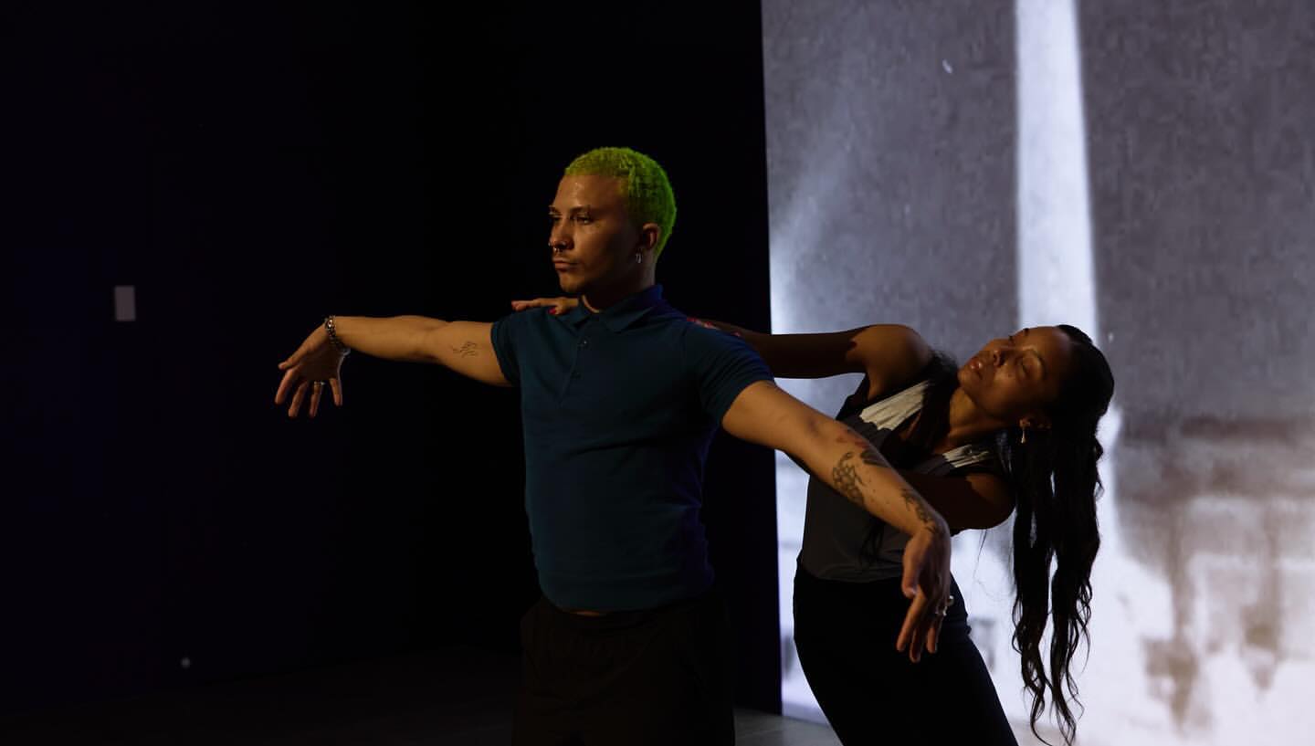 Two dancers, one male with green hair and one female, are captured mid-performance in a studio with dynamic lighting and choreography.
