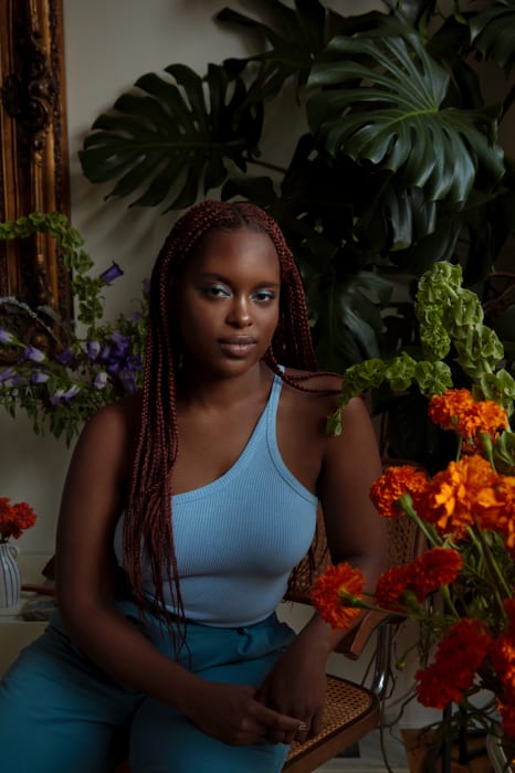 Portrait of a young woman with braided hair, wearing a light blue top, seated among lush green plants and colorful flowers indoors.