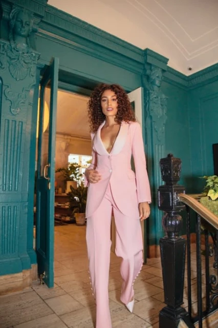 A brunette woman with curly hair poses in a pink suit with white heels in a brightly lit teal and white hallway.