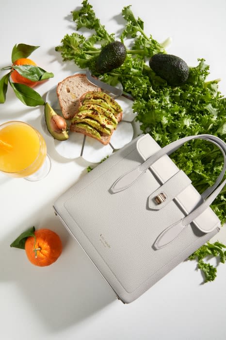 Grey leather handbag surrounded by fresh produce, including oranges, avocados, and leafy greens, presented against a white background.