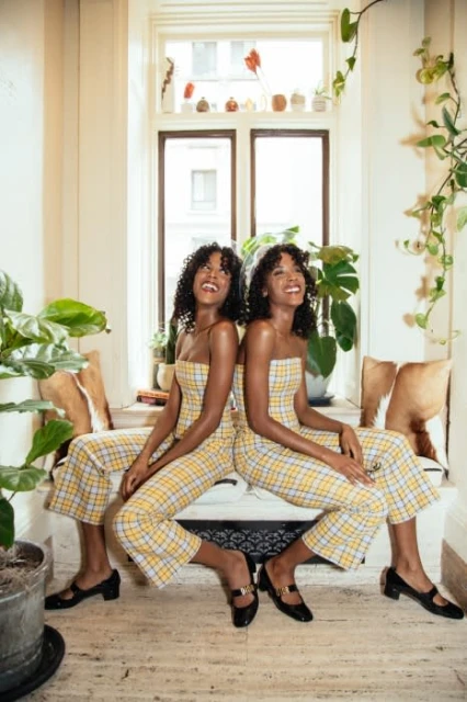 Two smiling African-American women in yellow plaid jumpsuits sit back-to-back on an indoor bench flanked by plants in a sunlit room.