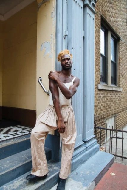 A Black man in modern avant-garde fashion poses on stairs outside a brick building, blending elegance and urban style in daylight.