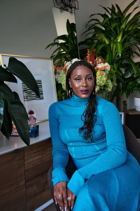 Portrait of an adult female in a blue dress sitting indoors. She has braided hair and is posed in front of plants and framed art.