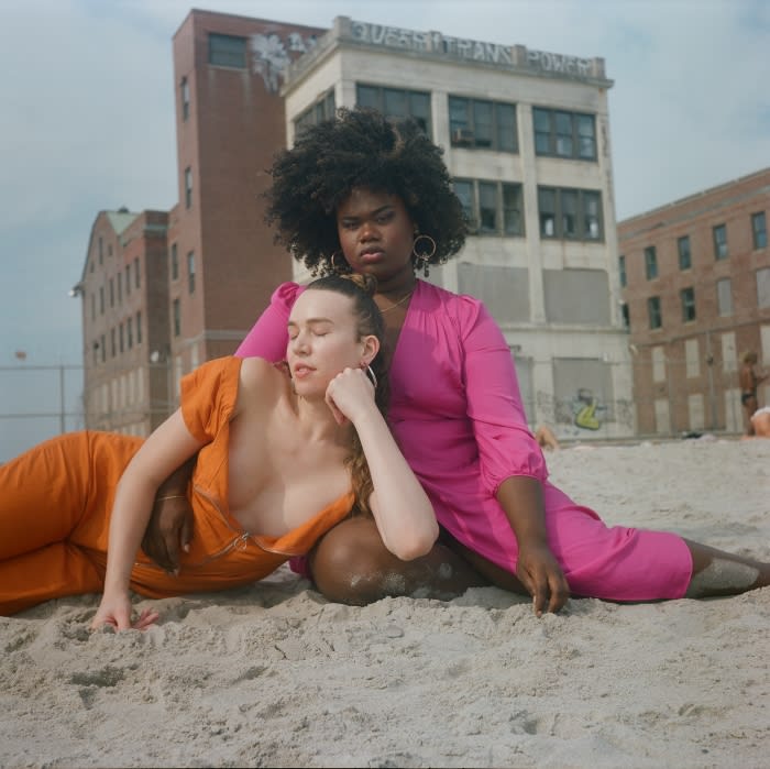 Two women in bright dresses pose on a sandy beach with brick buildings in the background under a bright daytime sky. 