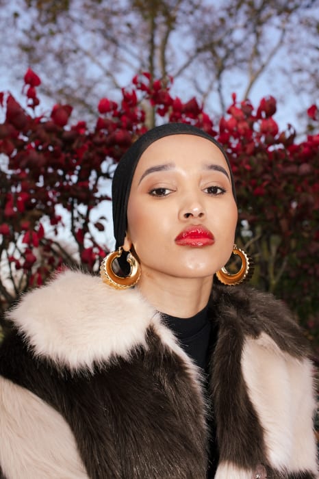 Close-up of a woman in a fur coat with hoop earrings against a red foliage backdrop, showcasing fashion and beauty elements.