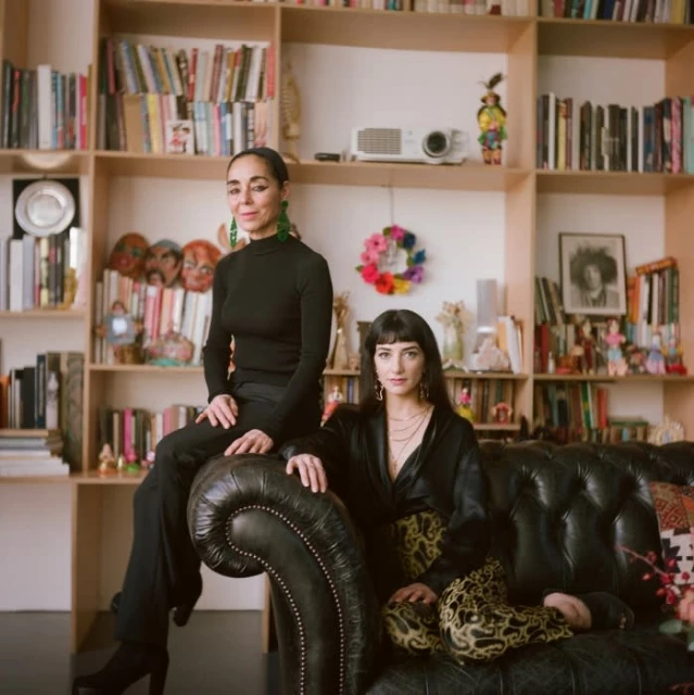 Two adult women in a home interior with bookshelves, sitting on a leather couch in front of a variety of home decor.