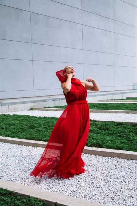 A joyful woman in a flowing red dress stands on a gravel path with a concrete wall and green ground cover in the background.