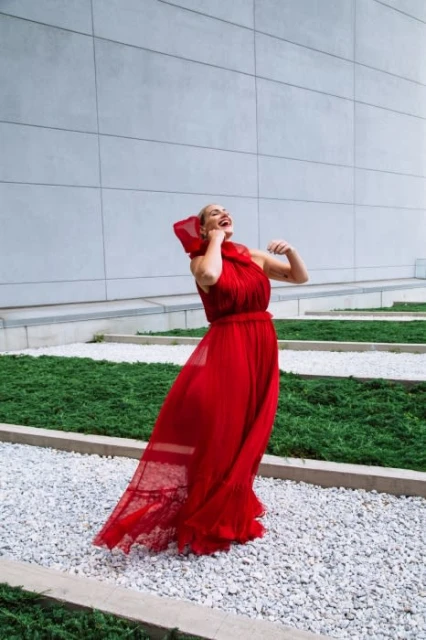 A joyful woman in a flowing red dress stands on a gravel path with a concrete wall and green ground cover in the background.