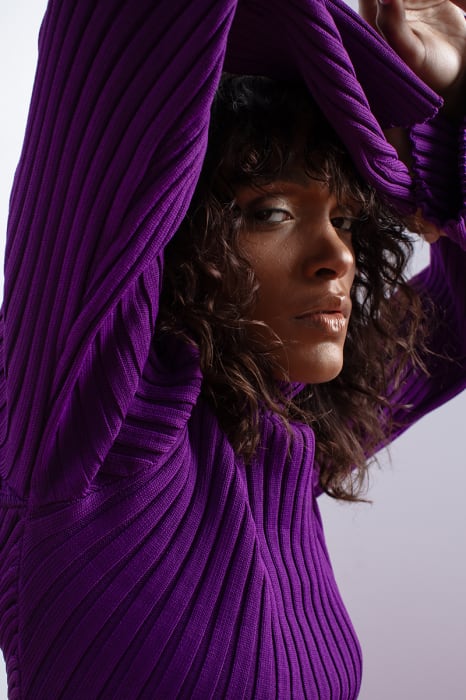 Dark-skinned woman in purple ribbed turtleneck sweater with hands above head, facing forward with natural makeup in a studio setting.