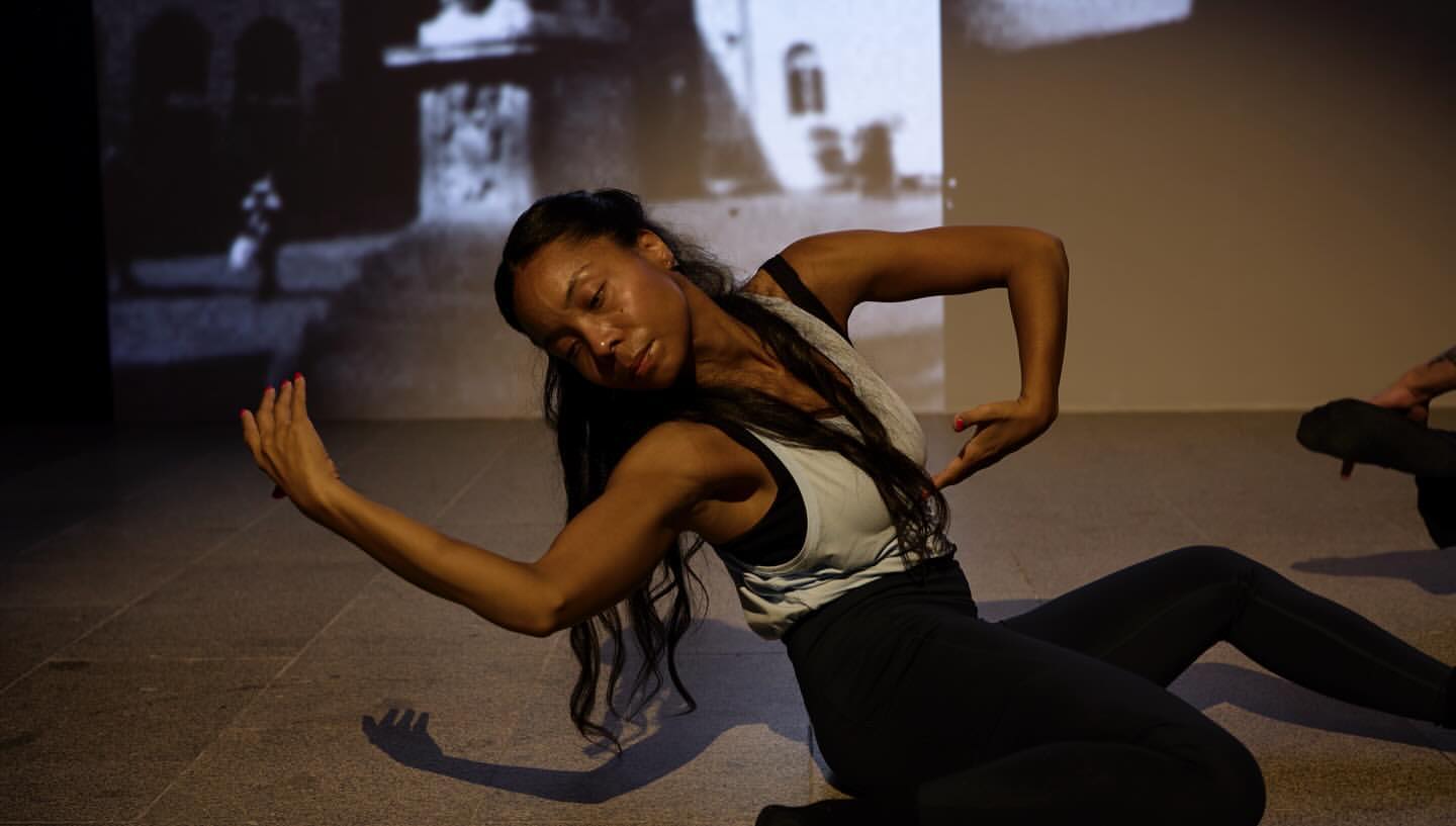 Black woman dancer performing a floor movement with extended arms against a projected backdrop