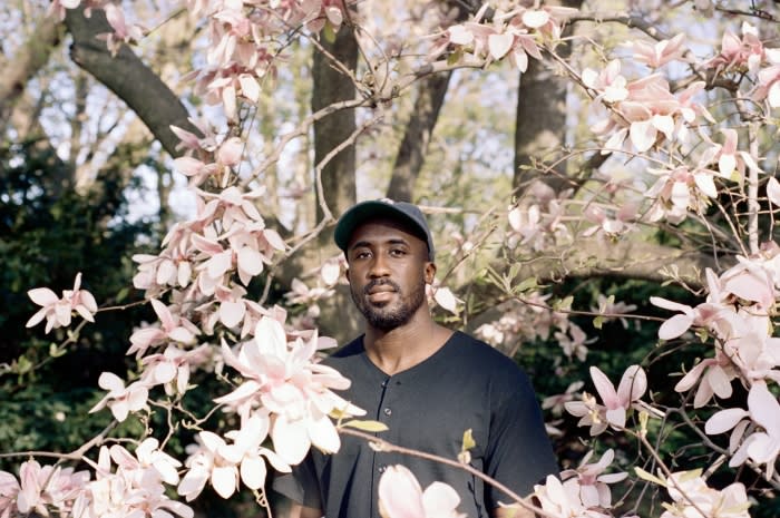 Adult male surrounded by blooming flowers in a close-up outdoor shot, wearing a hat and dark shirt, with trees in the background.