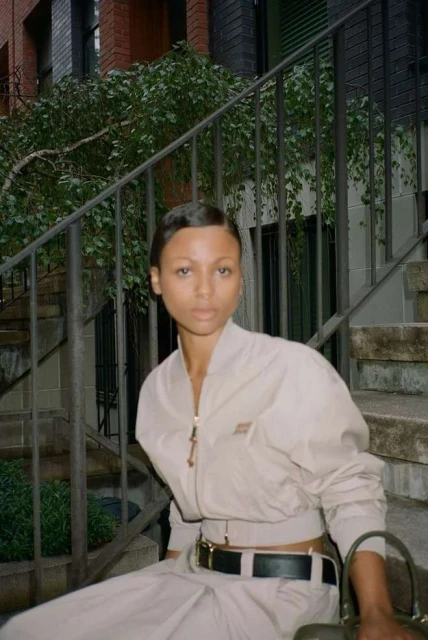 A dark-skinned woman with short hair sits on stairs wearing a tan jacket and pants, holding a green handbag, in an urban setting.