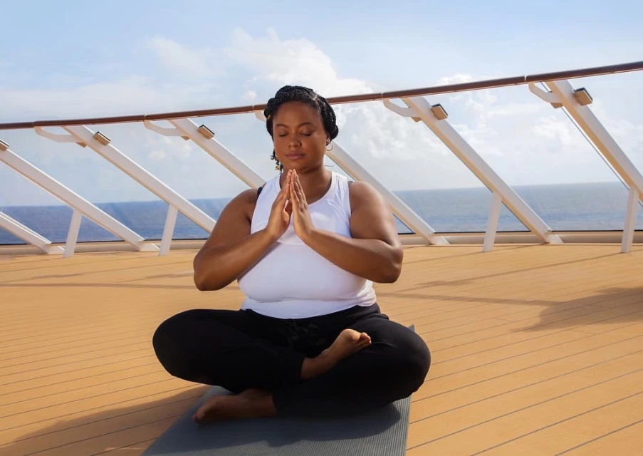 A woman meditates in lotus position on a yoga mat on the deck of a cruise ship, overlooking the ocean under a sunny sky.